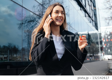 Smiling Woman in Business Attire Talks on the Phone While Walking Near a Modern Building in Bright Daylight Smiling Woman in Business Attire Talks on the Phone While Walking Near a Modern Building in Bright Daylight 132142617