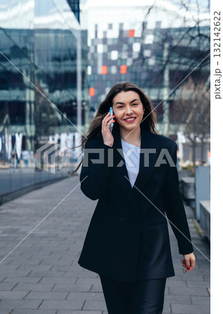 Professional Woman Walking Down City Sidewalk While Talking on Her Smartphone, Dressed in Stylish Black Blazer and Casual White Shirt on a Sunny Day Professional Woman Walking Down City Sidewalk While Talking on Her Smartphone, Dressed in Stylish Black Blazer and Casual White Shirt on a Sunny Day 132142622