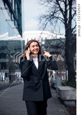 Businesswoman Walking in Urban Setting While Talking on Phone During Sunny Day Near Office Buildings 132142624