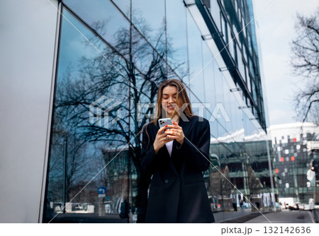 Young woman smiling while using smartphone outdoors near modern glass building reflecting trees and sky showcasing urban lifestyle concept 132142636