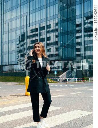 Confident young woman in a chic black suit smiles while speaking on her smartphone near a contemporary office building, showcasing a professional lifestyle Confident young woman in a chic black suit smiles while speaking on her smartphone near a contemporary office building, showcasing a professional lifestyle 132142644