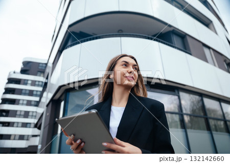 Confident businesswoman holding tablet while standing outside modern building with sleek architecture showcasing urban lifestyle concept Confident businesswoman holding tablet while standing outside modern building with sleek architecture showcasing urban lifestyle concept 132142660