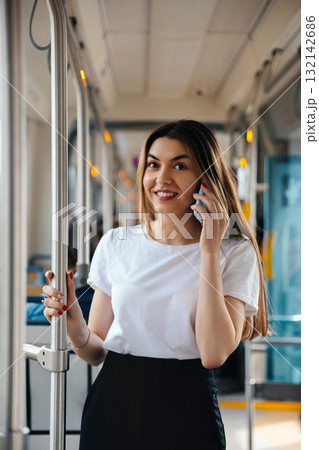 Young woman talking on smartphone while standing in public transport with a cheerful expression and stylish outfit showcasing urban lifestyle concept 132142686