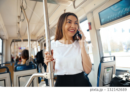 Young woman smiling while talking on smartphone in public transport setting with passengers around her showcasing urban lifestyle concept Young woman smiling while talking on smartphone in public transport setting with passengers around her showcasing urban lifestyle concept 132142689