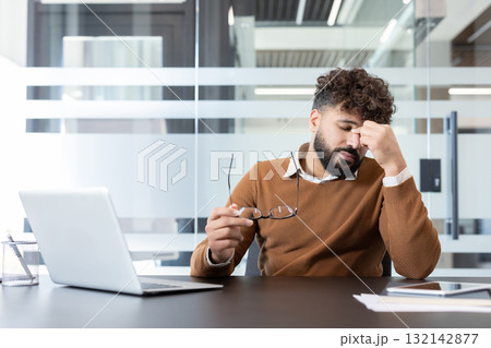Young man dealing with work burnout and headache, holding spectacles while sitting at office desk with laptop, experiencing exhaustion and stress from demanding workload Young man dealing with work burnout and headache, holding spectacles while sitting at office desk with laptop, experiencing exhaustion and stress from demanding workload 132142877
