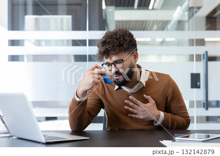 Young man experiencing an asthma attack and struggling with breathing difficulty at his workplace, using a blue inhaler while holding his chest with concern Young man experiencing an asthma attack and struggling with breathing difficulty at his workplace, using a blue inhaler while holding his chest with concern 132142879