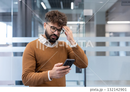 Man with a bearded face and eyeglasses holding a smartphone, experiencing headache and stress while reading bad news or difficult information in a modern office 132142901
