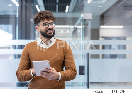 Young ethnic professional in smart-casual attire stands in a bright modern office, holding a tablet, smiling and looking away with confident, optimistic business energy 132142902