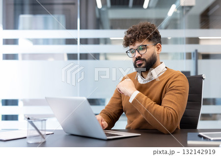 Young focused businessman working with a laptop at his desk in a blurred modern office environment, representing concentration, dedication, and professional productivity Young focused businessman working with a laptop at his desk in a blurred modern office environment, representing concentration, dedication, and professional productivity 132142919