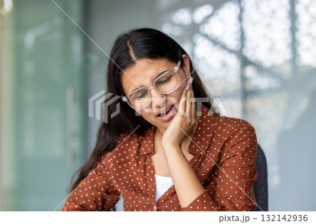 Businesswoman sitting at her desk in the office, experiencing a strong toothache, touching her cheek with her hand, reflecting discomfort and frustration while trying to focus on work Businesswoman sitting at her desk in the office, experiencing a strong toothache, touching her cheek with her hand, reflecting discomfort and frustration while trying to focus on work 132142936