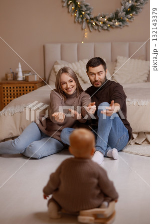 Young parents playing with baby on floor in cozy bedroom with beige wall and Christmas wreath Young parents playing with baby on floor in cozy bedroom with beige wall and Christmas wreath 132143379
