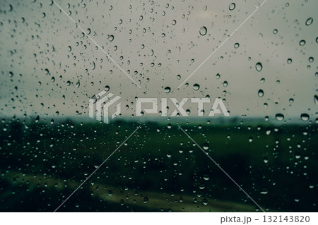 Raindrops on a wet glass window with blurred green landscape in the background. Moody rainy day, nature melancholy, peaceful atmosphere, seasonal weather and emotional calmness. Autumn fall season. 132143820