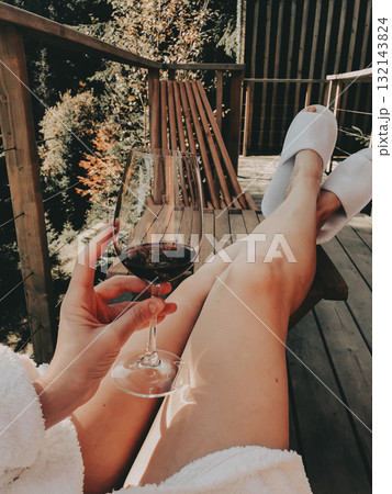 Relaxing moment on a wooden terrace with a glass of red wine in hand, legs in cozy slippers stretched out, enjoying calm nature views and warm sunlight in a peaceful outdoor setting. Travel girl. 132143824