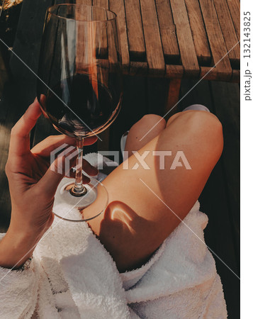 Relaxing moment on a wooden terrace with a glass of red wine in hand, legs in cozy slippers stretched out, enjoying calm nature views and warm sunlight in a peaceful outdoor setting. Travel girl. 132143825