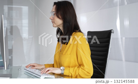 Businesswoman wearing yellow casual blouse and glasses is typing on laptop at office glass desk, analyzing data for business tasks. Audit in business Businesswoman wearing yellow casual blouse and glasses is typing on laptop at office glass desk, analyzing data for business tasks. Audit in business 132145588