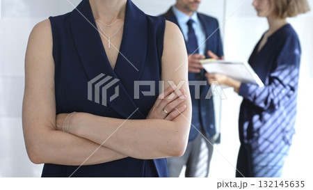 Professional businesswoman stands confidently with arms crossed in sharp focus as colleagues interact behind her during an office meeting, symbolizing leadership and corporate success 132145635