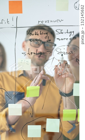 Business colleagues brainstorming, writing strategy notes on glass board, collaborating with colorful sticky notes in creative workplace meeting Business colleagues brainstorming, writing strategy notes on glass board, collaborating with colorful sticky notes in creative workplace meeting 132145682