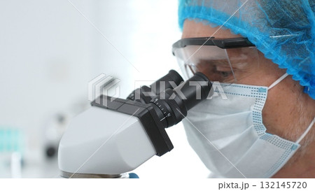 Masked scientist examining sample through microscope, wearing protective lab gear during medical research. Medicine, healthcare and science concept 132145720