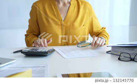 Female accountant wearing yellow shirt is examining a financial report with a magnifying glass while using a calculator and laptop in an office setting, close up. Audit and taxes in business Female accountant wearing yellow shirt is examining a financial report with a magnifying glass while using a calculator and laptop in an office setting, close up. Audit and taxes in business 132145770