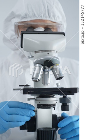 Male virologist in a hazmat suit, mask, and blue gloves, diligently working at a microscope in a laboratory researching viruses. Medicine, science concept 132145777
