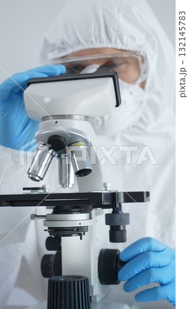 Male virologist in a hazmat suit, mask, and blue gloves, diligently working at a microscope in a laboratory researching viruses. Medicine, science concept 132145783