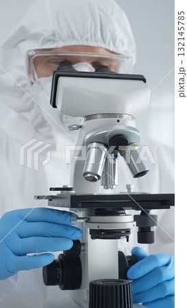 Male virologist in a hazmat suit, mask, and blue gloves, diligently working at a microscope in a laboratory researching viruses. Medicine, science concept 132145785