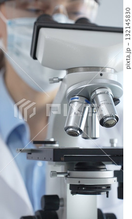 Scientist wearing lab coat and protective glasses using a modern microscope in a laboratory setting, conducting research and experiments in a scientific environment. Health care and science concept Scientist wearing lab coat and protective glasses using a modern microscope in a laboratory setting, conducting research and experiments in a scientific environment. Health care and science concept 132145830