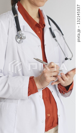 Close up of woman doctor wearing stethoscope and white coat over brick red blouse is standing and writing on a clipboard, representing medical expertise and patient care. Medicine concept Close up of woman doctor wearing stethoscope and white coat over brick red blouse is standing and writing on a clipboard, representing medical expertise and patient care. Medicine concept 132145837