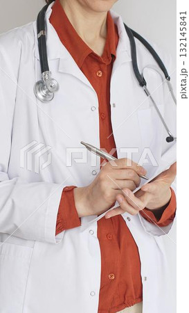 Close up of woman doctor wearing stethoscope and white coat over brick red blouse is standing and writing on a clipboard, representing medical expertise and patient care. Medicine concept 132145841