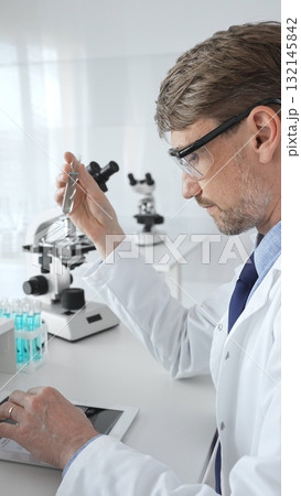 Male researcher in protective glasses holding test tube with blue liquid and working with tablet in sterile laboratory environment. Medicine and science concept 132145842