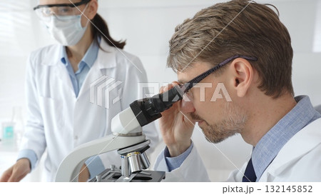 Scientific professional examining specimen through microscope, laboratory assistant nearby wearing protective mask, both dressed in white lab coats during research process. Medicine and science 132145852