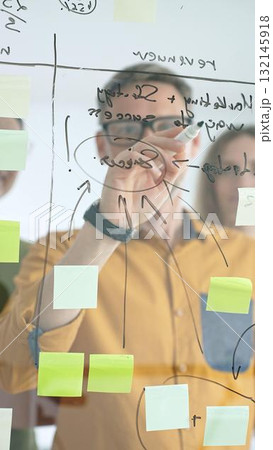 Business colleagues brainstorming, writing strategy notes on glass board, collaborating with colorful sticky notes in creative workplace meeting 132145918
