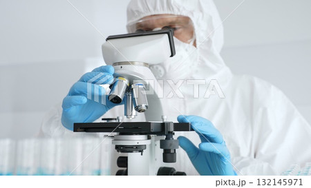 Male scientific researcher or virologist wearing protective gear adjusting microscope, analyzing lab samples with precision in sterile research environment. Medicine concept 132145971