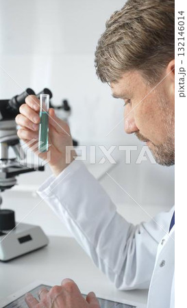 Senior male scientific researcher wearing white lab coat, analyzing blue liquid sample with tablet and microscope during medical investigation. Medicine and science concept 132146014