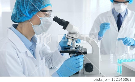Woman scientist in blue protective gear is examining specimen under microscope while laboratory assistant handles glassware in medical investigation at the background. Medicine, and science concept Woman scientist in blue protective gear is examining specimen under microscope while laboratory assistant handles glassware in medical investigation at the background. Medicine, and science concept 132146062