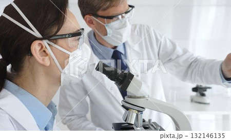 Two scientists in lab coats and protective gear conduct research in a sterile laboratory: one observes through a microscope, and the other holds a test tube with turquoise liquid. Medicine and science Two scientists in lab coats and protective gear conduct research in a sterile laboratory: one observes through a microscope, and the other holds a test tube with turquoise liquid. Medicine and science 132146135