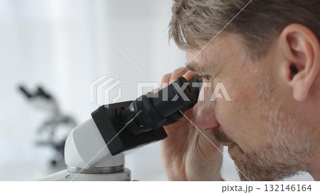 Close up of a man scientist carefully observing through a microscope in a brightly lit laboratory, conducting research for developing new medicines and treatments. Medicine and science concept Close up of a man scientist carefully observing through a microscope in a brightly lit laboratory, conducting research for developing new medicines and treatments. Medicine and science concept 132146164