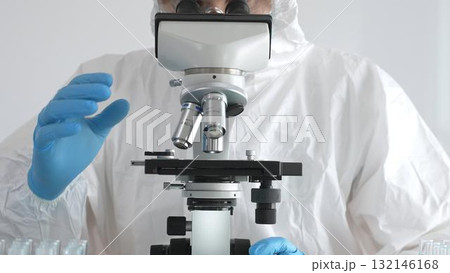 Medical virologist in protective hazmat gear examining viral samples under a microscope while conducting research in a sterile laboratory environment 132146168