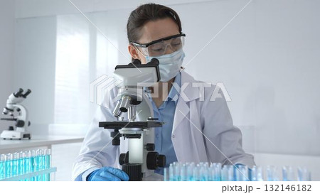 A female scientist in a lab coat and blue gloves examines a microscope slide in a sterile laboratory, conducting research using blue samples from a test tube A female scientist in a lab coat and blue gloves examines a microscope slide in a sterile laboratory, conducting research using blue samples from a test tube 132146182