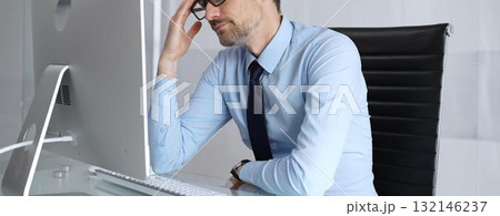 Overworked businessman experiencing headache and painful migraine, feeling overwhelming stress and burnout while working long hours at computer in a modern office setup 132146237