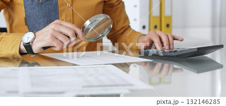 Accountant analyzing financial data with magnifying glass, calculating expenses, and auditing documents at office desk. Audit and taxes in business Accountant analyzing financial data with magnifying glass, calculating expenses, and auditing documents at office desk. Audit and taxes in business 132146285
