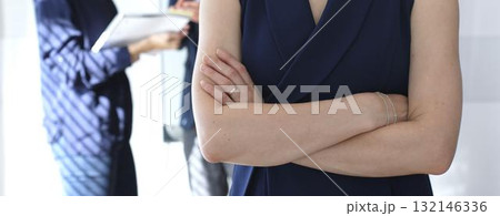 Professional businesswoman stands confidently with arms crossed in sharp focus as colleagues interact behind her during an office meeting, symbolizing leadership and corporate success 132146336