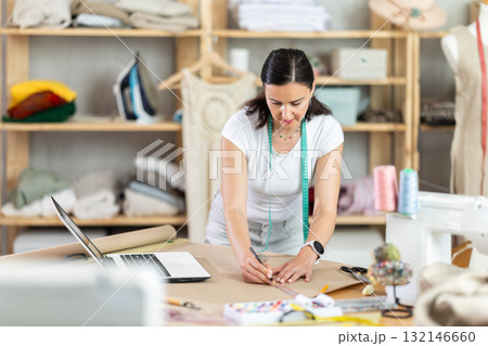 Woman tailor draws dress pattern of product on paper, laptop on table. 132146660