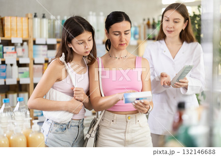 Attentive mother and daughter buyers choosing medical ointment and professional female parmacist helping in pharmacy 132146688