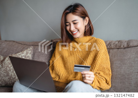 A smiling young woman in a sweater sits on a sofa with a credit card in hand, making an online purchase using a laptop. Concept of Black Friday and online shopping 132147482