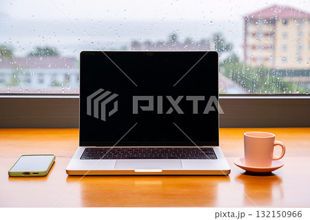 Laptop with blank screen, smartphone and coffee cup on wooden table. Remote work setup on rainy day. 132150966