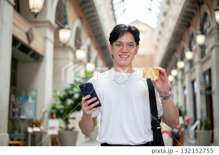 Smiling asian man with glasses in white tee holding phone and showing credit card in shopping mall Smiling asian man with glasses in white tee holding phone and showing credit card in shopping mall 132152755