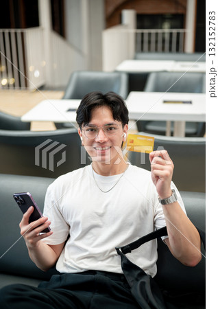 Asian man wearing glasses in white tee holding phone and showing his credit card while sit on sofa. 132152783