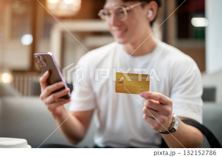 Asian man wearing glasses in white shirt is holding smartphone and credit card while sitting in cafe 132152786