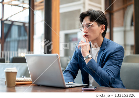 Blue suit asian man thinking thoughtfully in front of a laptop while sitting in cafe or coffee shop. Blue suit asian man thinking thoughtfully in front of a laptop while sitting in cafe or coffee shop. 132152820